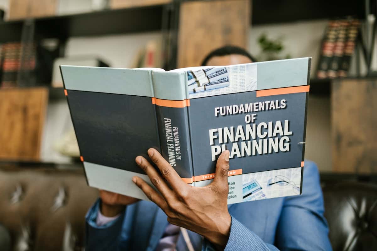 A man in a blue suit sitting on a couch reading a book called Fundamentals of Financial Planning.