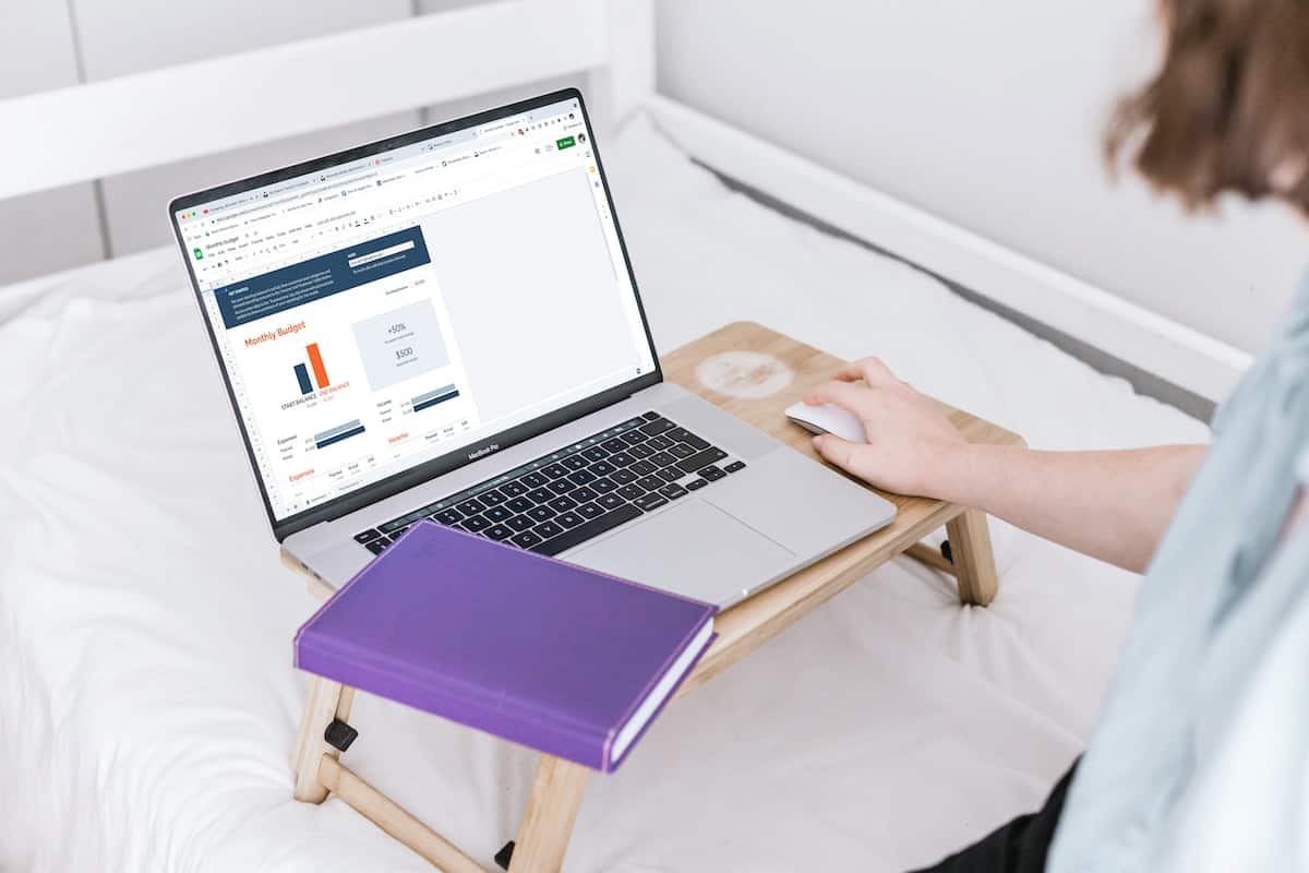 Person using a laptop in bed next to a purple book, with the laptop screen displaying a monthly budget chart.