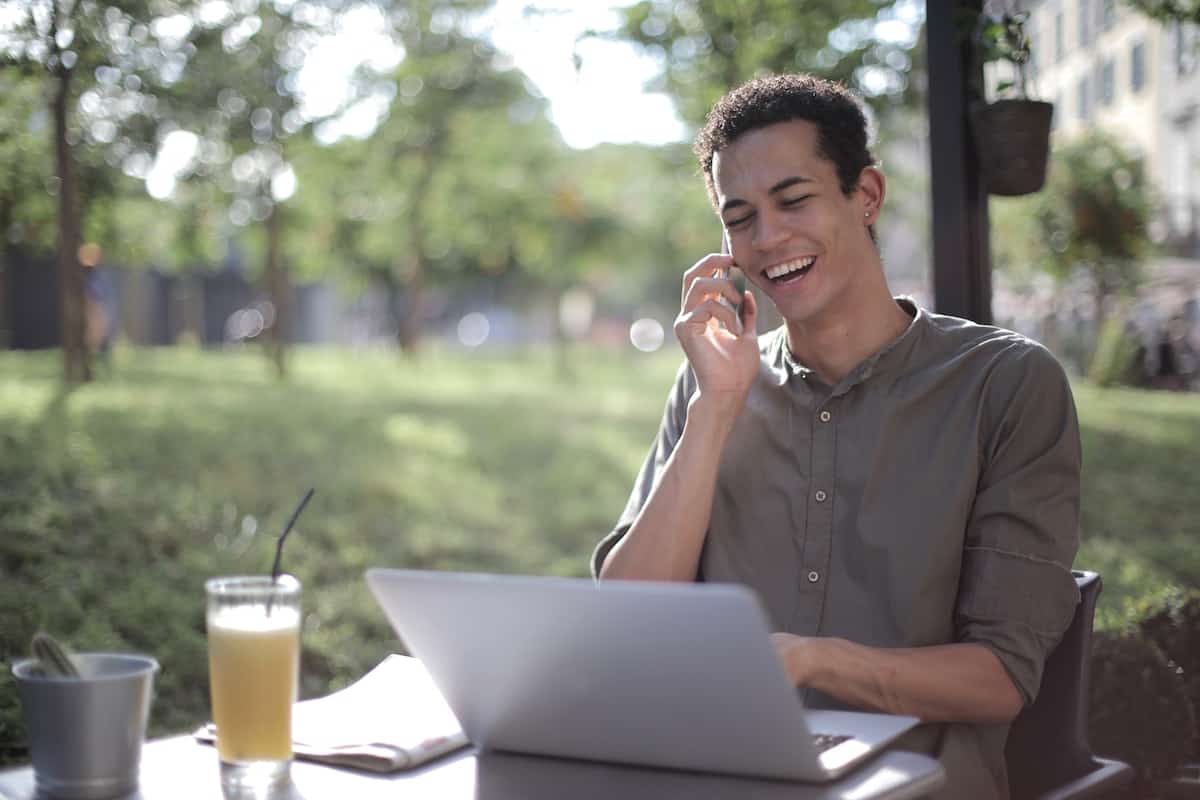 A man sitting at an outdoor table while talking on the phone and typing on a laptop.