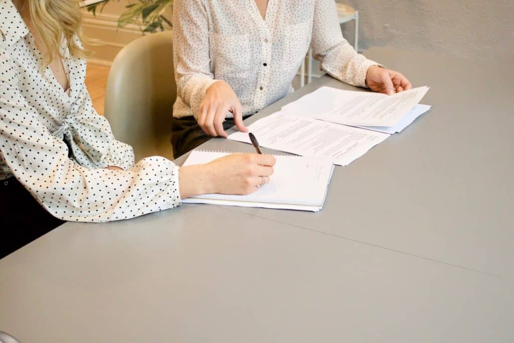 Two women facing each other at a gray table with pens and paper.
