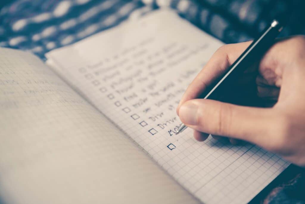 A hand holds a pen while writing a checklist into a notebook.