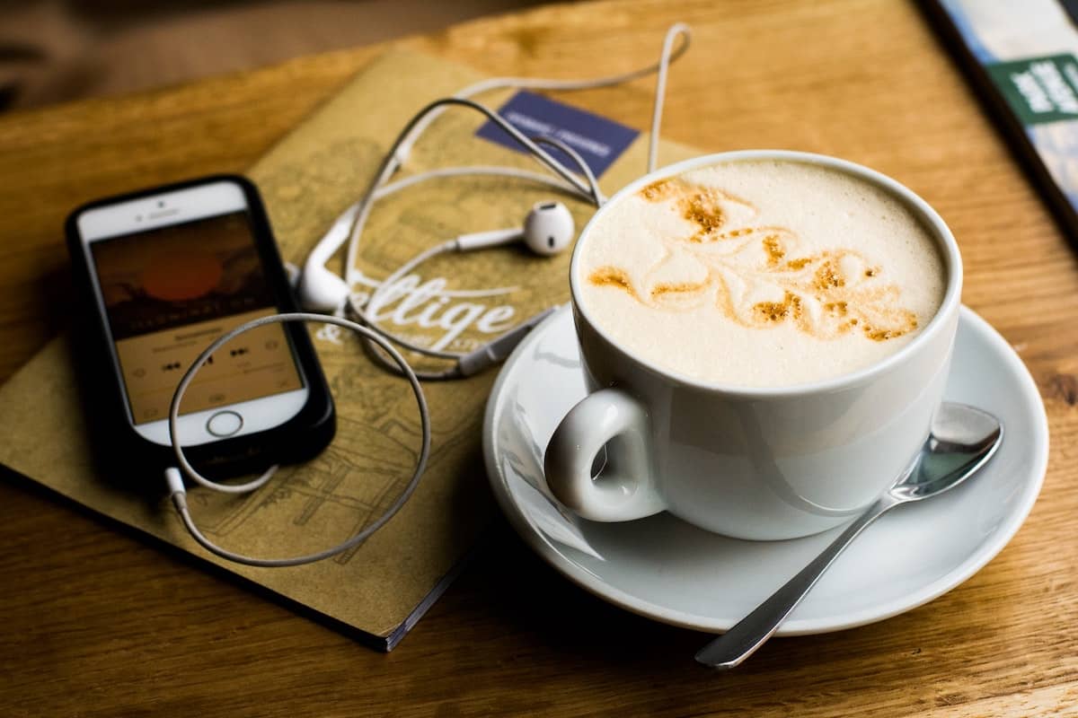 A cellphone playing a podcast next to a cup of coffee.