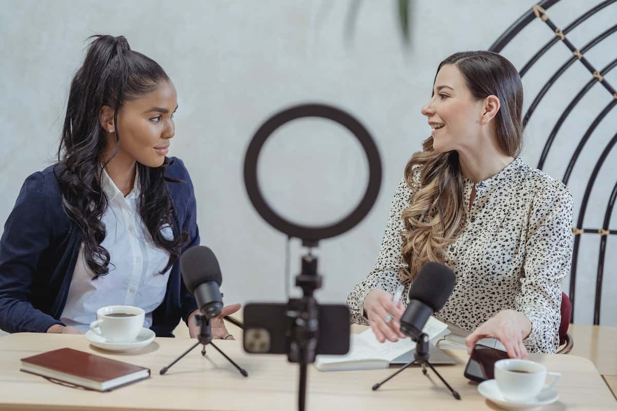 Two ladies sitting at a desk and recording a podcast show in front of a phone and ring light