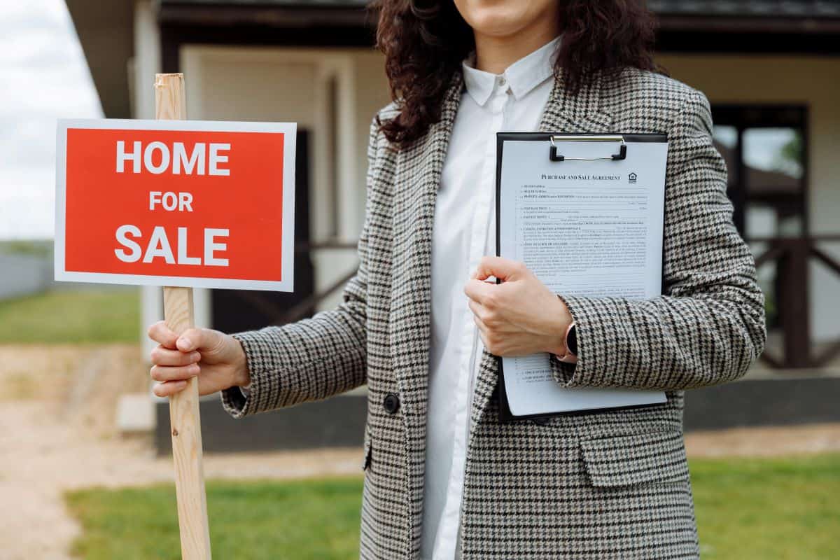 Realtor standing holding a clipboard and “For sale” sign, with a house in the background.
