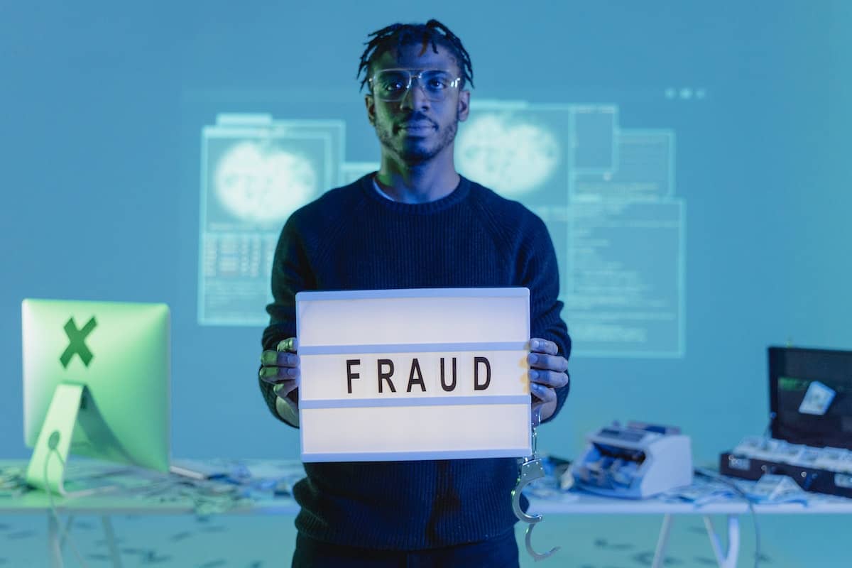 Man with glasses standing in front of a desk wearing a black sweater holding a letter lightbox, that says “fraud”